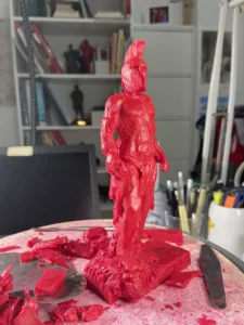 Figura esculpida en cera roja representando un guerrero clásico en proceso de modelado, sobre una mesa de trabajo.
